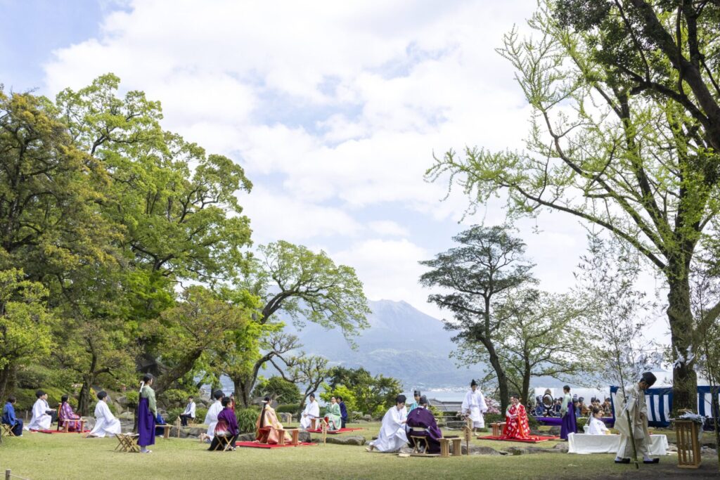 前回の曲水宴の開催風景。桜島を望む庭園で、伝統装束の参宴者たちが和歌を詠む優雅なひととき。